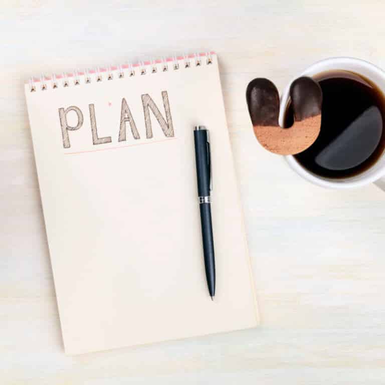 Lucky plan. An overhead photo of a spiral notebook with the handwritten word Plan, with a cup of coffee with a lucky horseshoe shaped cookie, on a desk with copy space