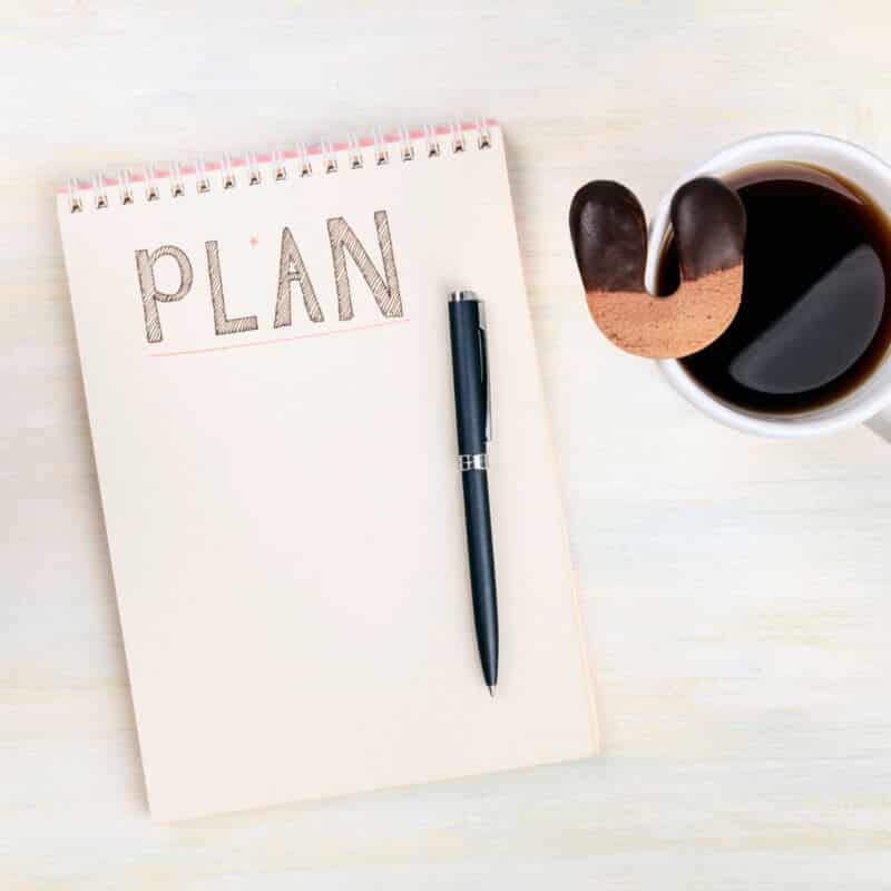 Lucky plan. An overhead photo of a spiral notebook with the handwritten word Plan, with a cup of coffee with a lucky horseshoe shaped cookie, on a desk with copy space
